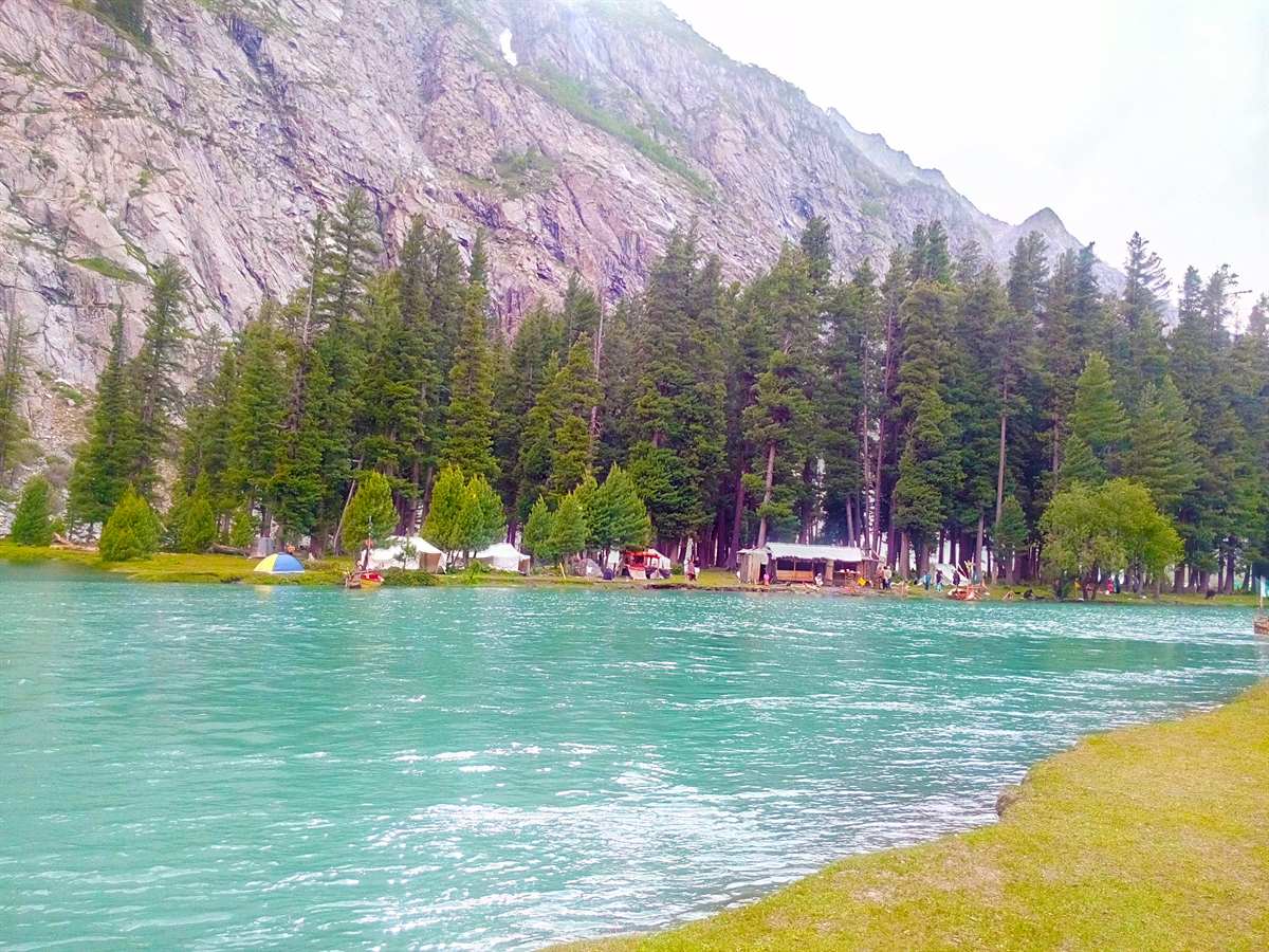 Turquoise water of Mahodand Lake with boats, lakeside tents and pine forest below rocky peaks