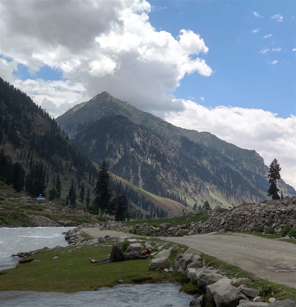 Green mountainside and the milky Gabral river in summer, with a local man napping on the riverside grass